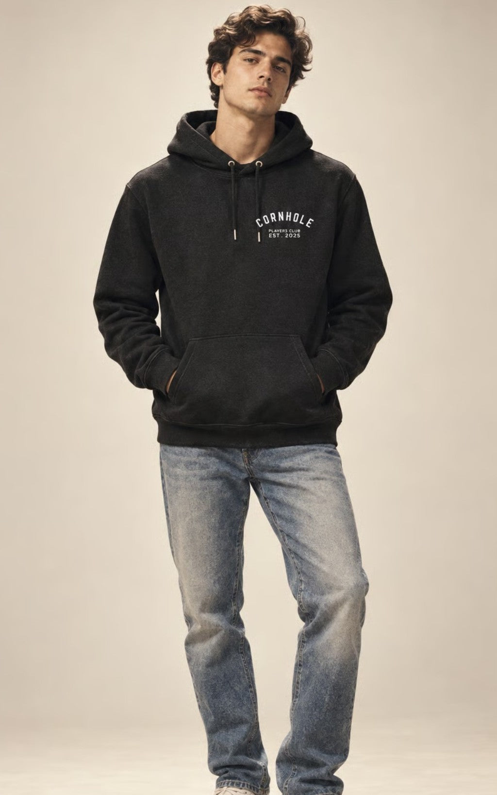 A man wearing a dark charcoal gray hoodie with 'CRONHOLE EST.2025' printed in white on the chest, paired with light-wash blue jeans, standing against a neutral beige studio background.